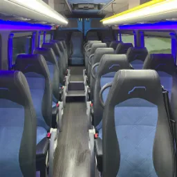 Wnętrze autobusu z rzędami czarno-niebieskich foteli z logo na zagłówkach, oświetlone niebieskim i żółtym światłem.