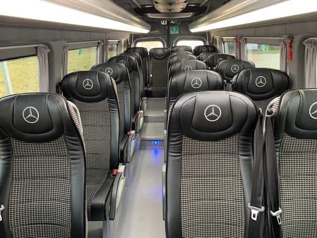 Wnętrze komfortowego busa Mercedes z rzędami foteli z logo marki na zagłówkach, widok z przodu pojazdu, oświetlenie sufitowe.