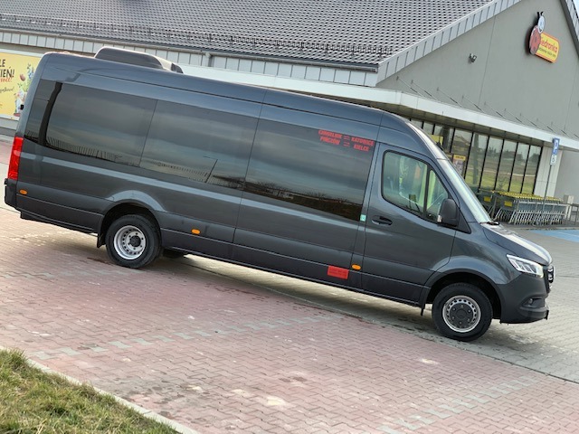 Szary bus Mercedes-Benz Sprinter z przyciemnianymi szybami, zaparkowany na chodniku przed sklepem spożywczym, z napisem na boku wymieniającym miasta: Chmielnik, Katowice, Pińczów, Kielce.
