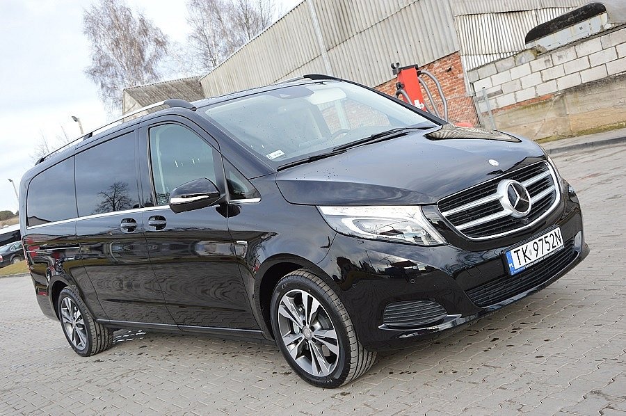 Czarny Mercedes-Benz V-klasa z relingami dachowymi, zaparkowany na kostce brukowej przed budynkiem gospodarczym.