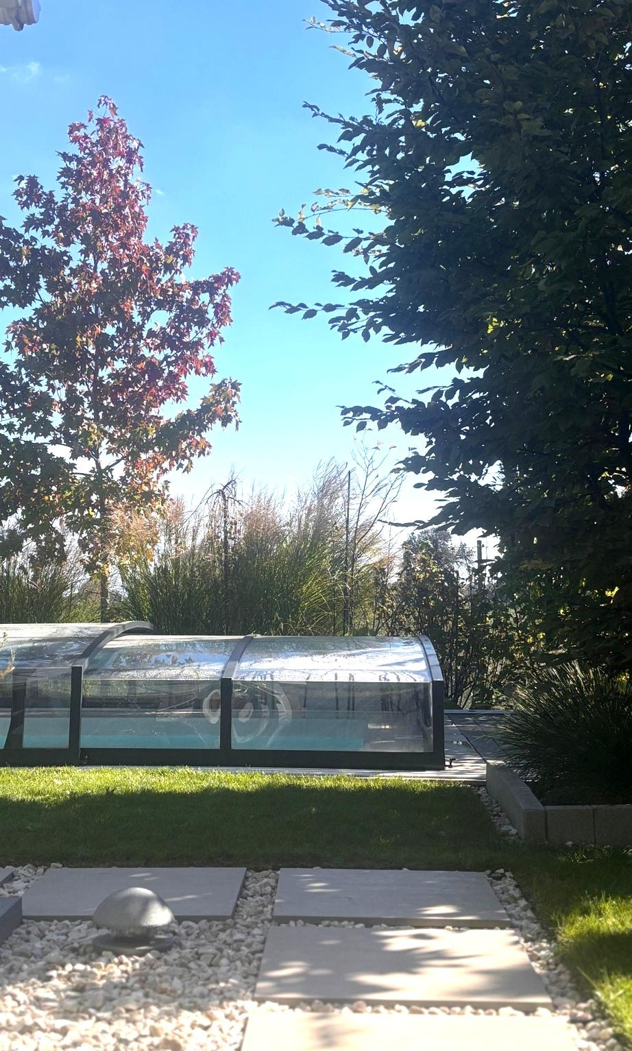 Ogród z basenem przy domu szeregowym: trawnik, ścieżka z płyt i kamieni, drzewa w tle. Basen zadaszony. Efekt po realizacji projektu.