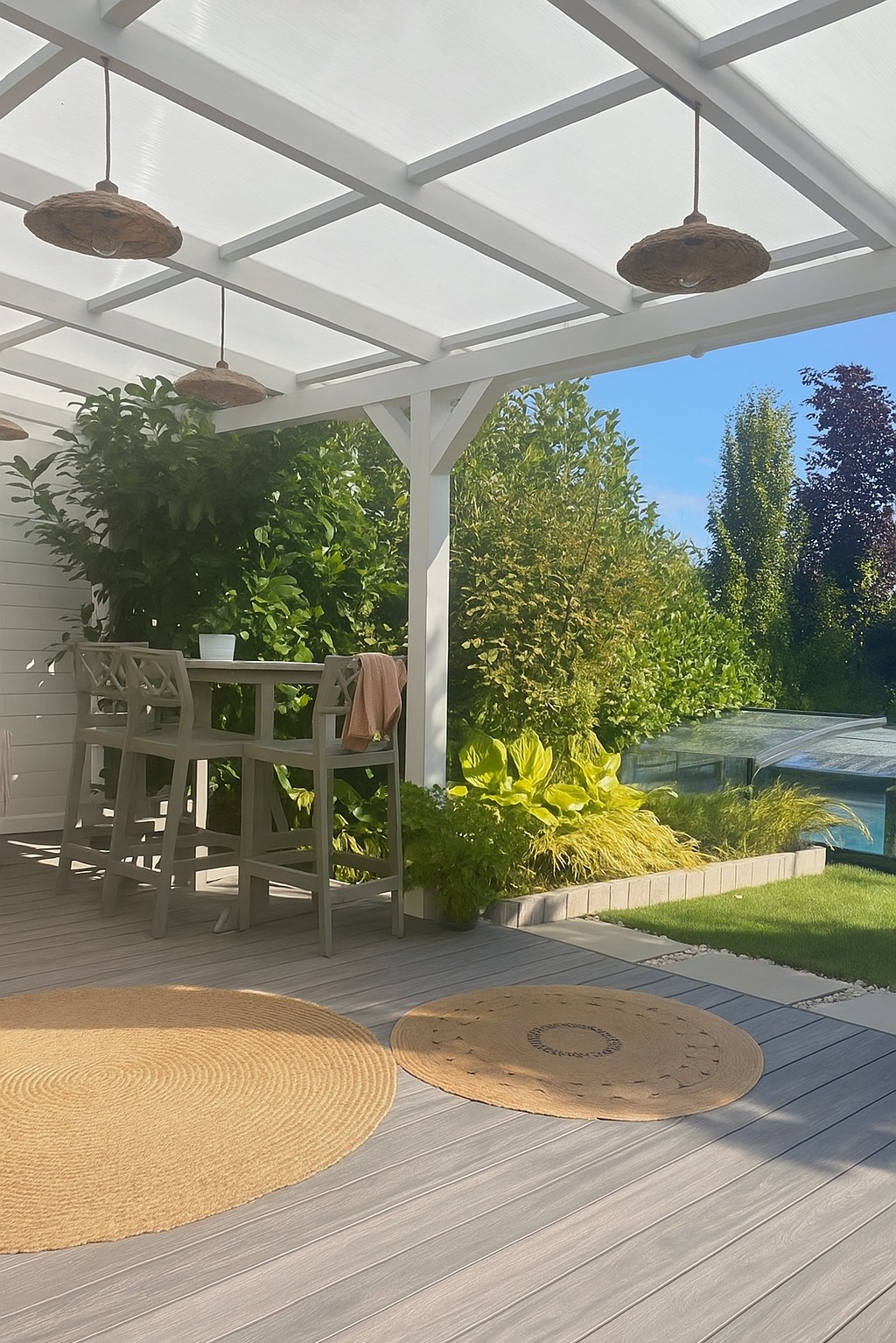 Zadaszony taras z drewnianą podłogą i okrągłymi dywanami, widok na zadbany ogród z basenem. Uwagę przykuwają lampy w stylu boho pod sufitem.