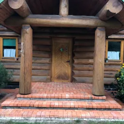 Wejście do drewnianego domu z bali, wyłożone czerwoną kostką brukową, schody z kostki, drewniane kolumny i drzwi.