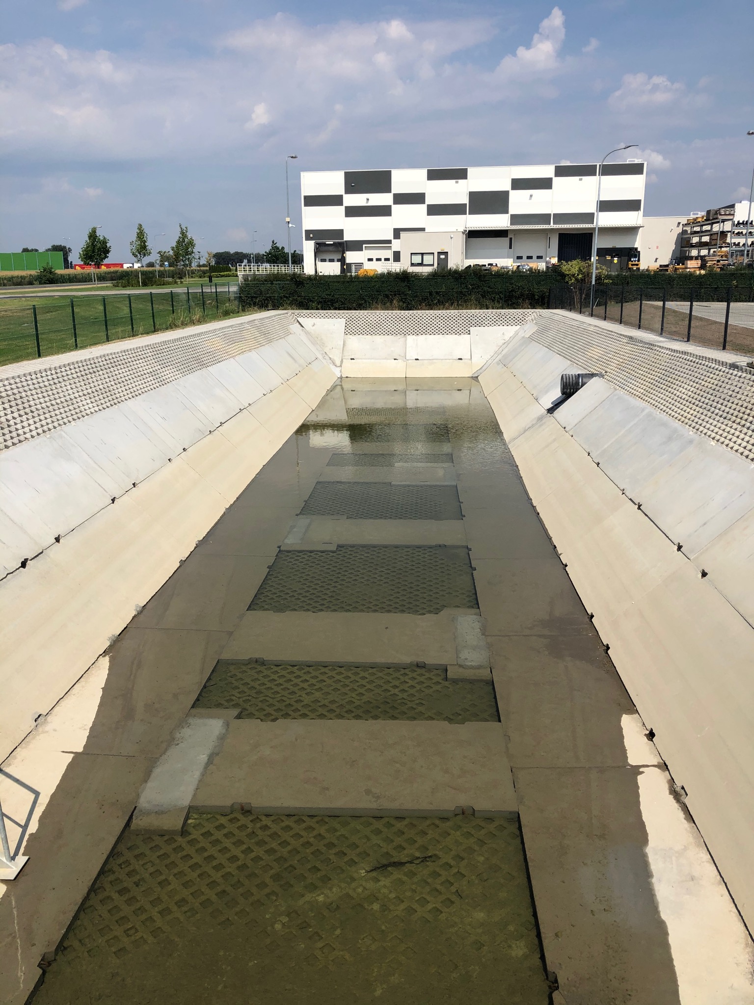 Betonowy kanał odwadniający z ażurowymi płytami na dnie, częściowo wypełniony wodą, na tle budynku przemysłowego w biało-czarne kwadraty.