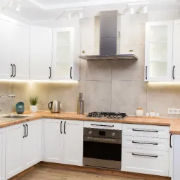 Nowoczesna, jasna kuchnia z białymi szafkami i drewnianym blatem, wyposażona w sprzęt AGD: piekarnik, płytę gazową, mikrofalówkę i okap.