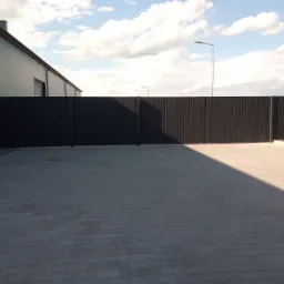 Długa, nowoczesna, metalowa brama wjazdowa w kolorze antracytowym, z pionowymi panelami, zamykająca wjazd do budynku przemysłowego, widoczna kostka brukowa i fragment pola w tle.