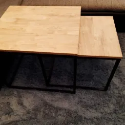 Dwa stoliki kawowe z drewnianymi blatami o jasnym odcieniu i czarnymi, metalowymi nogami, ustawione na puszystym, szarym dywanie, z fragmentem sofy w tle.