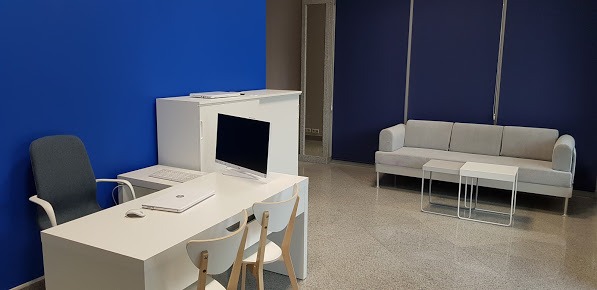 Jasne, nowoczesne biuro z białymi meblami, biurkiem z komputerem i laptopem, szarym krzesłem biurowym, dwoma białymi krzesłami i szarą sofą z dwoma stolikami kawowymi na tle niebieskich ścian.