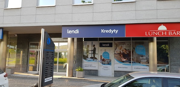 Fasada budynku z szyldem 'lendi Kredyty' obok 'Lunch Bar', z naklejkami na szybach prezentującymi sceny z klientami.