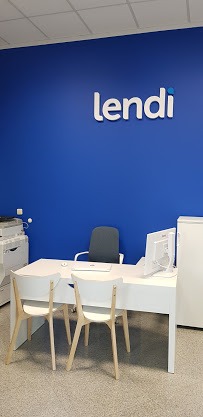Biuro z białym biurkiem, krzesłami, komputerem i logo 'lendi' na niebieskiej ścianie w tle.
