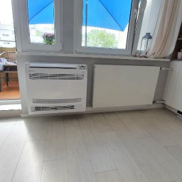 WOLTBERG Roman Mika - Biały klimatyzator podłogowy Rotenso obok grzejnika, na tle okna z widokiem na balkon i budynek, w jasnym wnętrzu z białymi meblami.