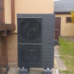 WOLTBERG Roman Mika - Szara pompa ciepła Panasonic Aquarea, ustawiona na betonowych blokach na żwirowym podłożu, przy beżowej ścianie budynku.