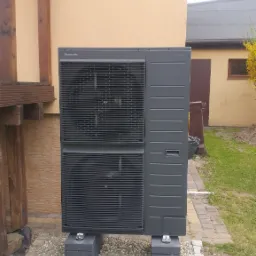 Szara pompa ciepła Panasonic Aquarea, ustawiona na betonowych blokach na żwirowym podłożu, przy beżowej ścianie budynku.