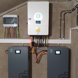 WOLTBERG Roman Mika - Kompletna instalacja fotowoltaiczna w pomieszczeniu: dwa szare magazyny energii na kółkach, biały inwerter Deye na ścianie i skrzynka z kablami. Widoczne kable i oświetlenie.