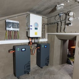 WOLTBERG Roman Mika - Kompletna instalacja magazynów energii w piwnicy z szarymi akumulatorami, białym inwerterem i licznikiem na ścianie. Widoczne kable i przewody elektryczne.