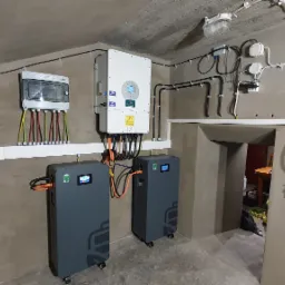 Kompletna instalacja magazyn&oacute;w energii w piwnicy z szarymi akumulatorami, białym inwerterem i licznikiem na ścianie. Widoczne kable i przewody elektryczne.