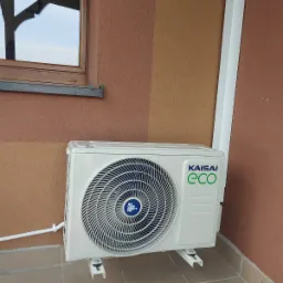 Biała jednostka zewnętrzna klimatyzacji Kaisai ECO zamontowana na brązowej elewacji budynku, widoczna z boku, z fragmentem okna w tle.