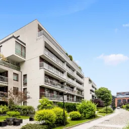 Nowoczesny kompleks apartamentowy z balkonami i zielenią, widok z poziomu chodnika z kostki brukowej, błękitne niebo.