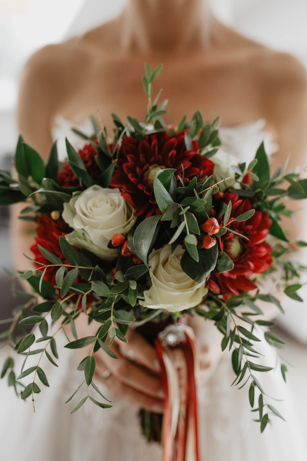 Panna młoda trzyma bukiet z białych róż, czerwonych dalii i zielonych liści eukaliptusa, przewiązany czerwonymi i białymi wstążkami.