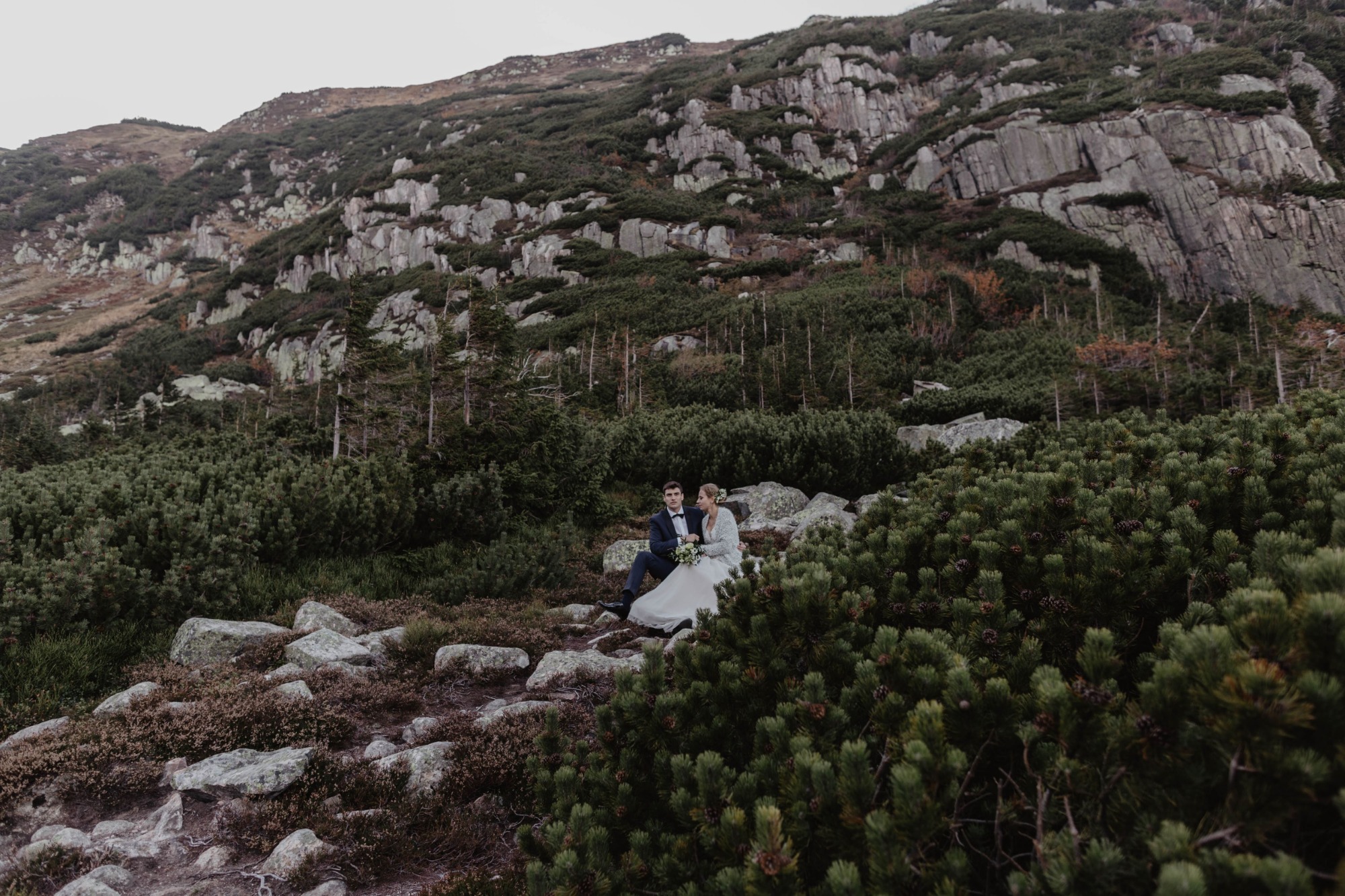 Para młoda siedzi na kamieniach w górskim krajobrazie, w tle skaliste zbocze porośnięte kosodrzewiną.