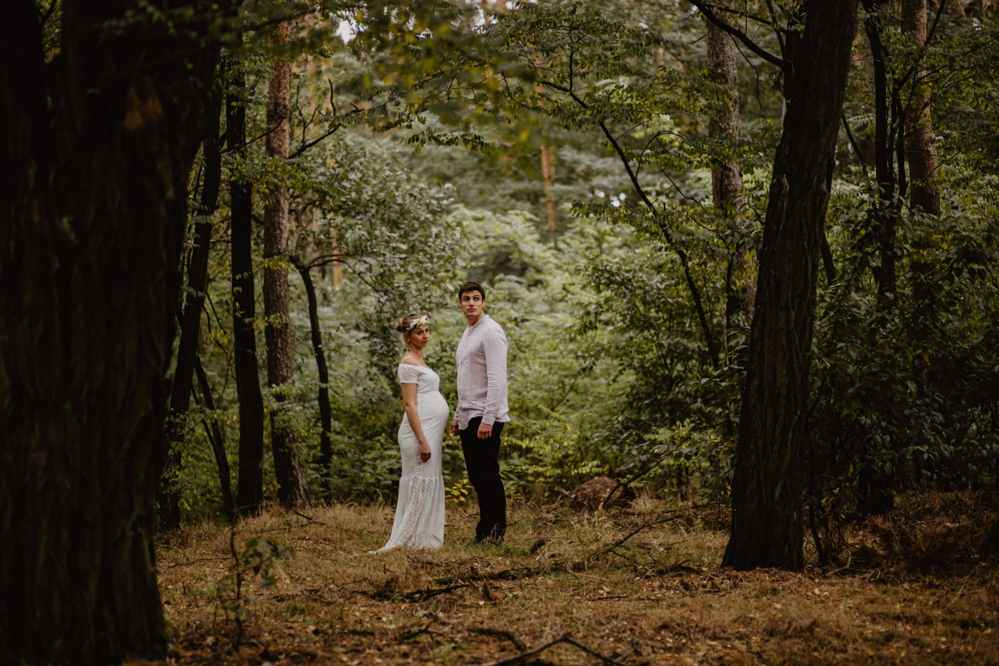 Ciężarna kobieta w białej koronkowej sukni i wianku na głowie, stoi obok mężczyzny w lesie, oboje patrzą w dal.