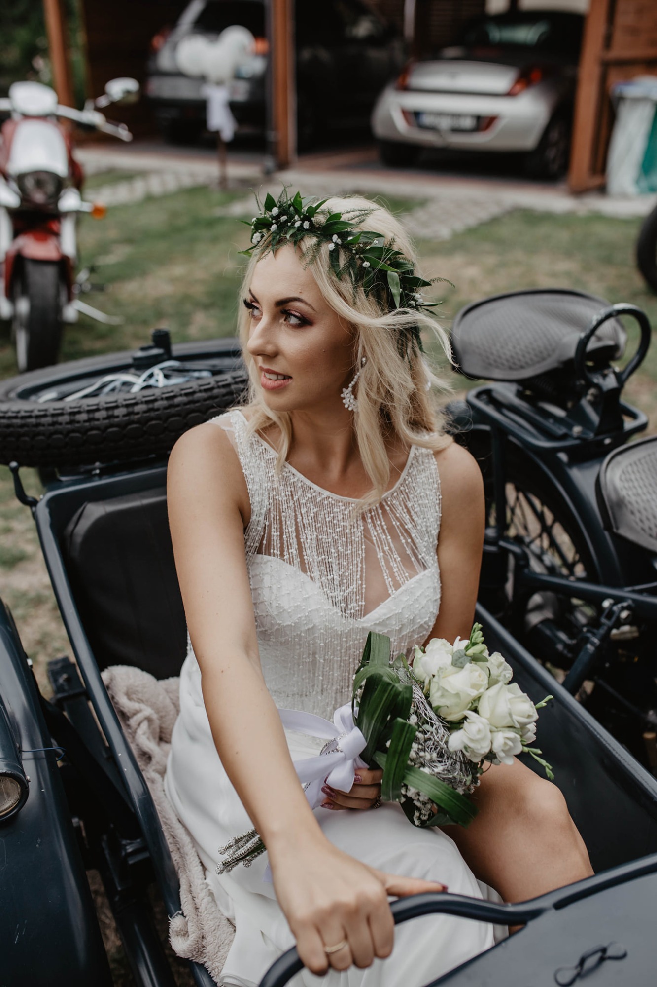 Panna młoda w białej sukni z wiankiem na głowie, siedząca w bocznym wózku motocykla z bukietem białych róż, spoglądająca w bok.