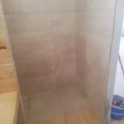 Wyłożona beżowymi płytkami kabina prysznicowa z drewnianą podłogą i widocznym niebieskim wiadrem z zaprawą w rogu.