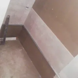 Ściana w trakcie przygotowywania do gładzi, widoczne profile, częściowo nałożona warstwa wyrównująca, narzędzia i materiały na podłodze, w tle brązowa ściana.