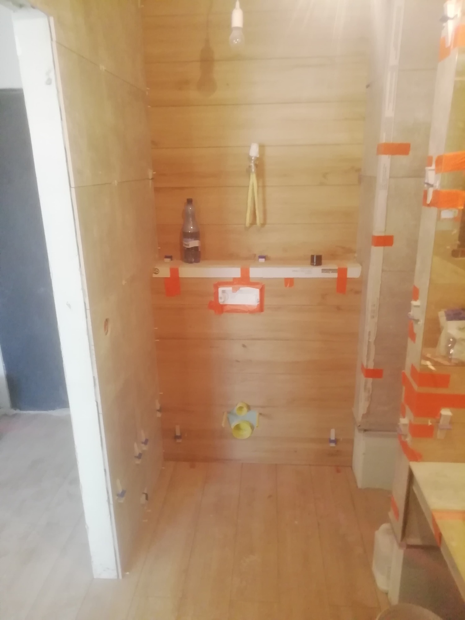 Łazienka w trakcie remontu: płytki imitujące drewno na ścianach i podłodze, poziomica z pomarańczową taśmą, butelka, żółta rura kanalizacyjna, widoczne spoiny płytek.