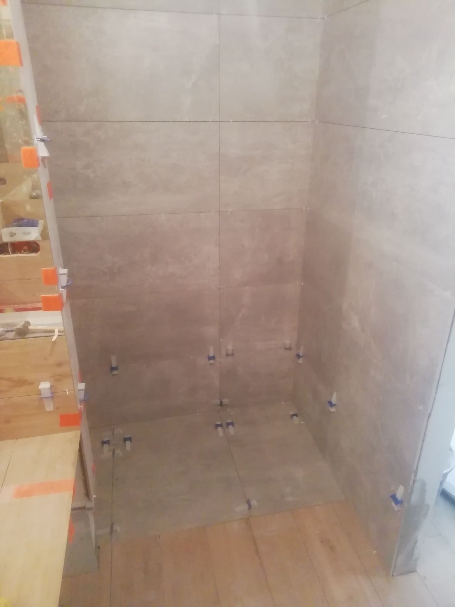 Ściany i podłoga w trakcie układania płytek ceramicznych w łazience, widoczne krzyżyki dystansowe i poziomica z pomarańczowymi elementami.