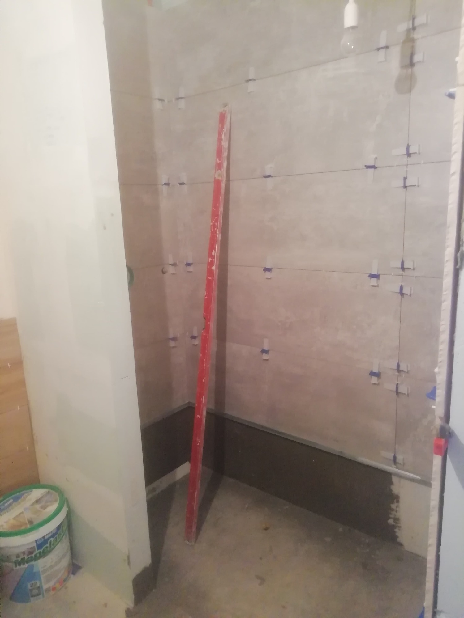 Ściana łazienki w trakcie układania płytek, widoczne sznurki wyznaczające linie, krzyżyki dystansowe, poziomica oparta o ścianę i wiadro z zaprawą klejową.