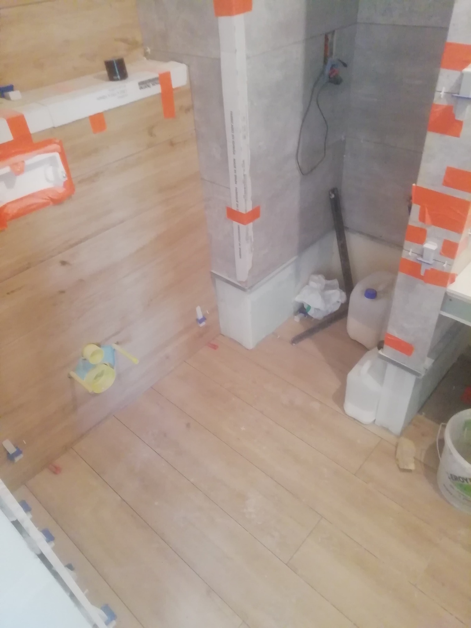 W trakcie układania płytek imitujących drewno na podłodze i ścianach w łazience, widoczne plastikowe elementy dystansowe, puszki instalacyjne i pojemniki z materiałami budowlanymi.