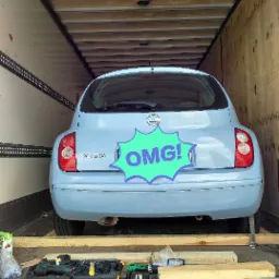 Niebieski Nissan Micra zabezpieczony drewnianymi belkami wewnątrz przestrzeni ładunkowej pojazdu transportowego, widoczne narzędzia i nap&oacute;j.