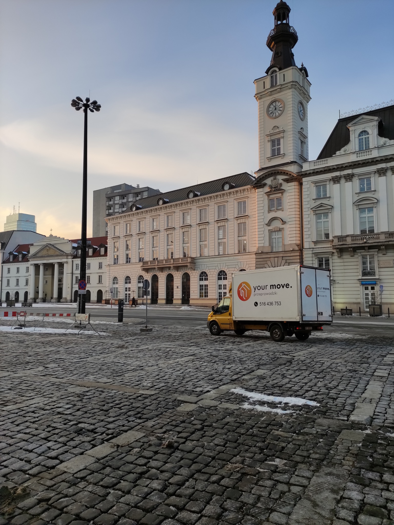 Żółty samochód dostawczy z logo firmy przeprowadzkowej na tle budynku z wieżą zegarową i brukowanego placu.
