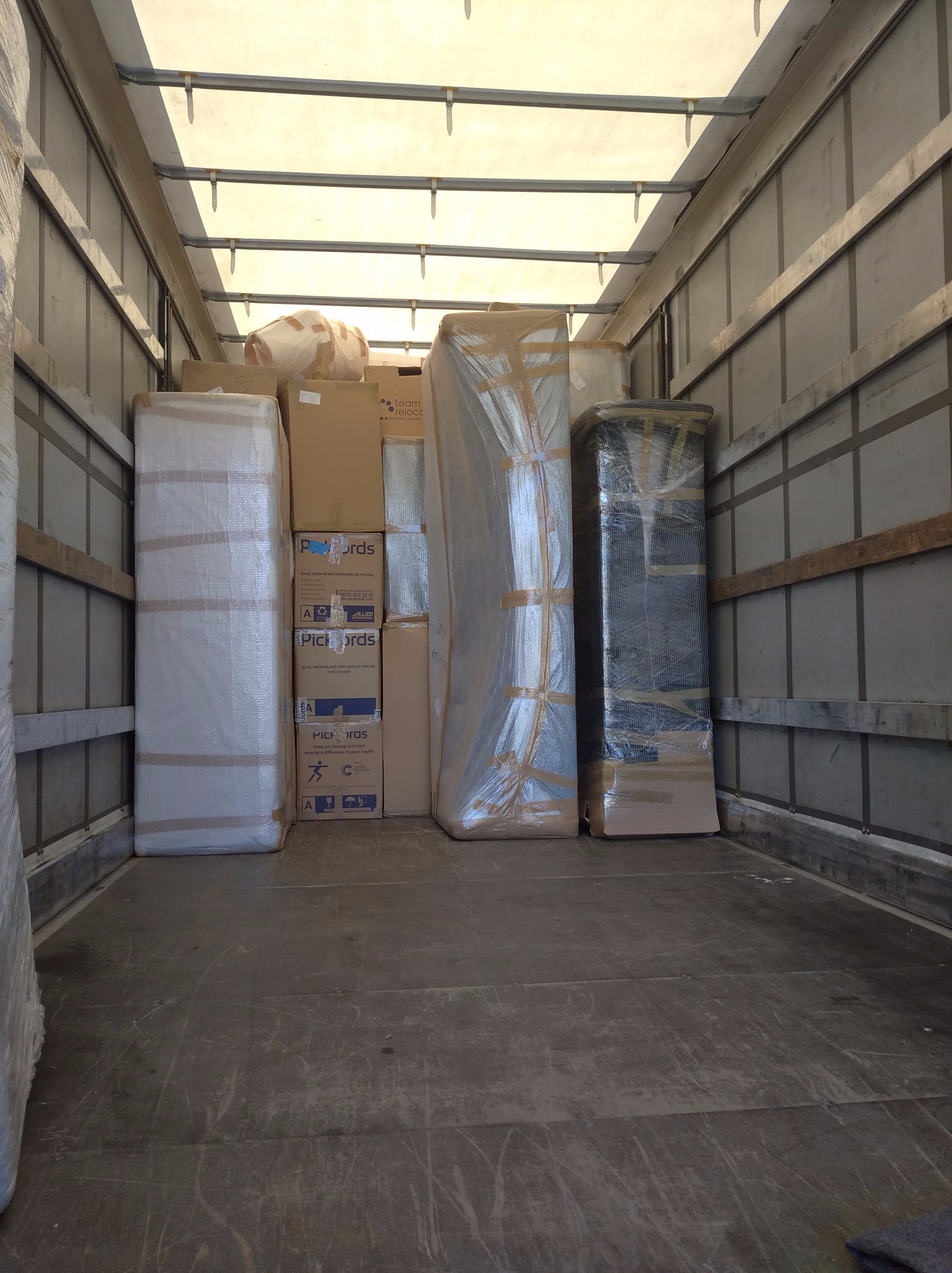 Wnętrze samochodu dostawczego wypełnione kartonami i meblami owiniętymi folią stretch, przygotowanymi do transportu. Widoczne logo 'Pickfords' na kilku kartonach.