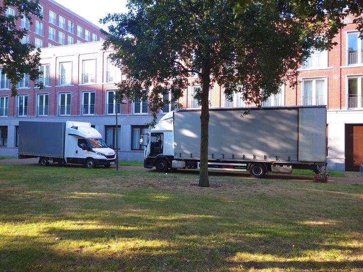 Dwa samochody dostawcze z plandekami zaparkowane na trawniku przed budynkiem mieszkalnym z cegły.