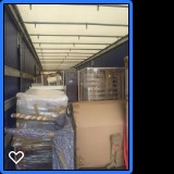 Wnętrze przestrzeni ładunkowej ciężarówki wypełnionej kartonami i meblami zabezpieczonymi folią stretch, widoczne oświetlenie sufitowe i niebieskie ściany boczne.