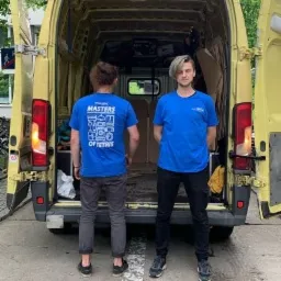 Dwaj mężczyźni w niebieskich koszulkach stoją przed otwartym, żółtym busem wypełnionym kartonami i torbami, jeden z nich ma blond włosy.