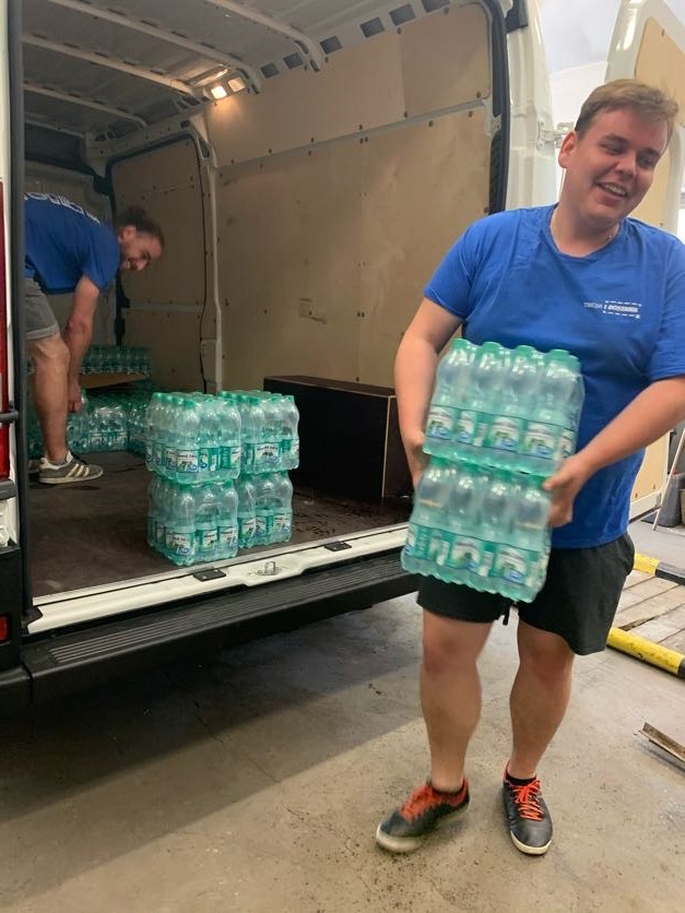 Dwóch mężczyzn w niebieskich koszulkach rozładowuje palety z wodą butelkowaną z białego furgonetki, jeden z nich uśmiecha się trzymając pakiet w rękach.