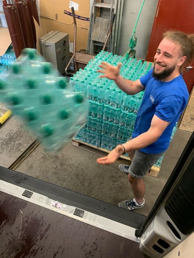 Uśmiechnięty mężczyzna w niebieskiej koszulce przenosi z palety do busa w Krakowie zgrzewkę butelek wody mineralnej, widoczna sterta palet z wodą.