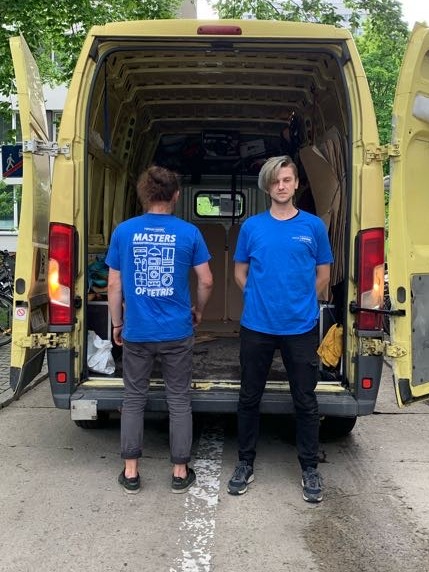 Dwaj mężczyźni w niebieskich koszulkach stoją przed otwartym, żółtym busem wypełnionym kartonami i torbami, jeden z nich ma blond włosy.