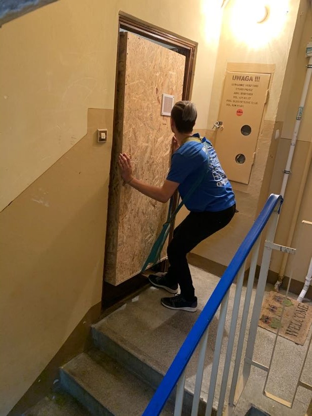 Mężczyzna w niebieskiej koszulce i czarnych spodniach, zabezpieczony pasami, ostrożnie przenosi duży, drewniany panel po schodach wewnątrz budynku. W tle widoczna kartonowa paczka z napisem 'UWAGA!