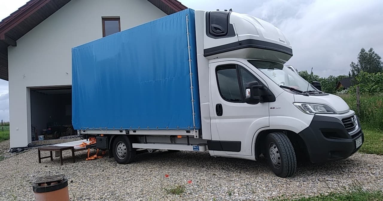 Biały bus z niebieską plandeką zaparkowany na żwirowym podjeździe przed białym domem z otwartym garażem, w tle zieleń.