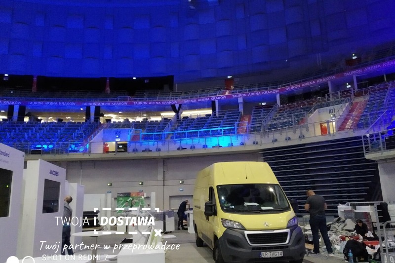 Żółty bus dostawczy Peugeot zaparkowany wewnątrz Tauron Areny Kraków podczas montażu stoisk targowych, widoczne logo firmy transportowej 'Twoja i Dostawa' na zdjęciu.