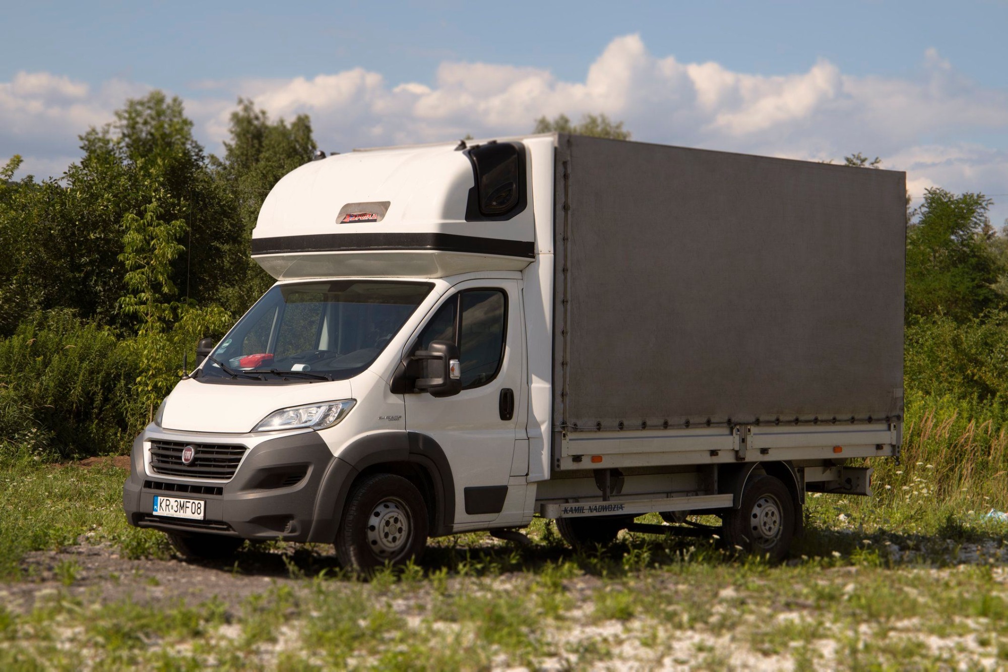 Biały samochód dostawczy Fiat Ducato z plandeką na tle zieleni i błękitnego nieba z chmurami.