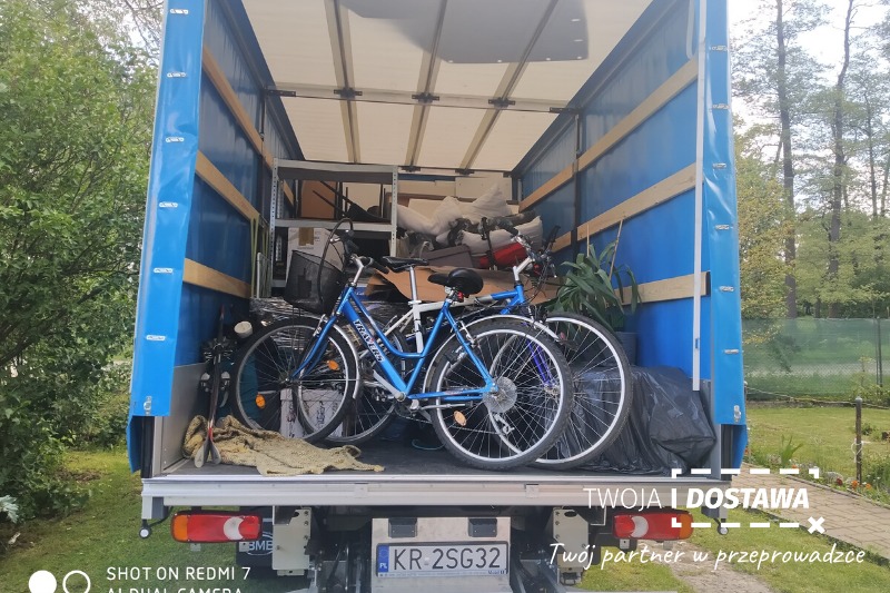 Wnętrze niebieskiej plandeki samochodu dostawczego wypełnione przedmiotami, w tym dwoma rowerami, kartonami i rośliną doniczkową, widoczne tablice rejestracyjne KR 2SG32.