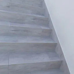 Schody zewnętrzne wyłożone jasnoszarą płytką imitującą drewno, z widoczną strukturą usłojenia, przylegające do białej ściany.