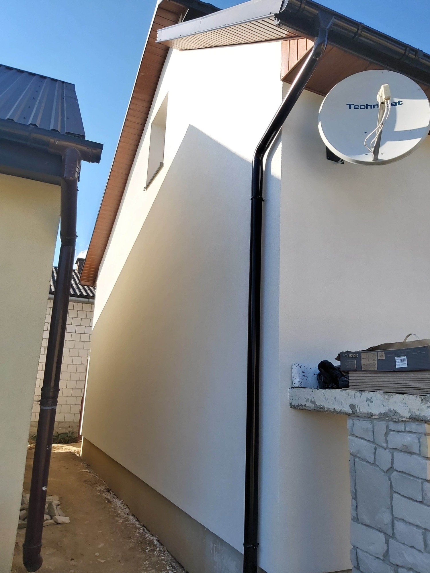 Fragment elewacji budynku z anteną satelitarną Technisat, widoczne orynnowanie, wąskie przejście między budynkami i materiały budowlane na podwyższeniu.