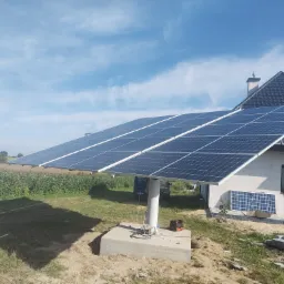 Wolnostojąca instalacja fotowoltaiczna z modułami skierowanymi w stronę nieba, umieszczona na betonowym fundamencie obok domu jednorodzinnego z ciemną dachówką, widoczne narzędzia i skrzynka...