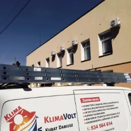 Biały samochód dostawczy z logo firmy Klimavolt i numerem telefonu na tle budynku z zamontowanymi klimatyzatorami i drabiną na dachu.